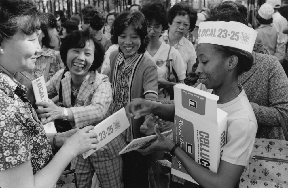 2-7-Corky-Lee-photo-Dudu-Masegale-organizer-at-Columbus-Park-rally-1982-giving-out-hats-scaled-img