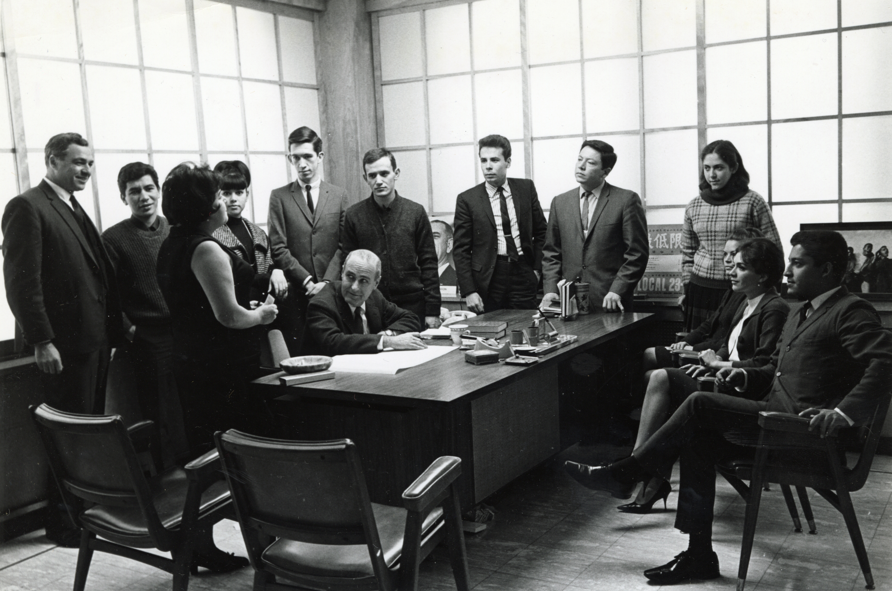 Kathy Andrade speaking to a group of young men and women gathered around a desk, with one older man seated at the desk listening.