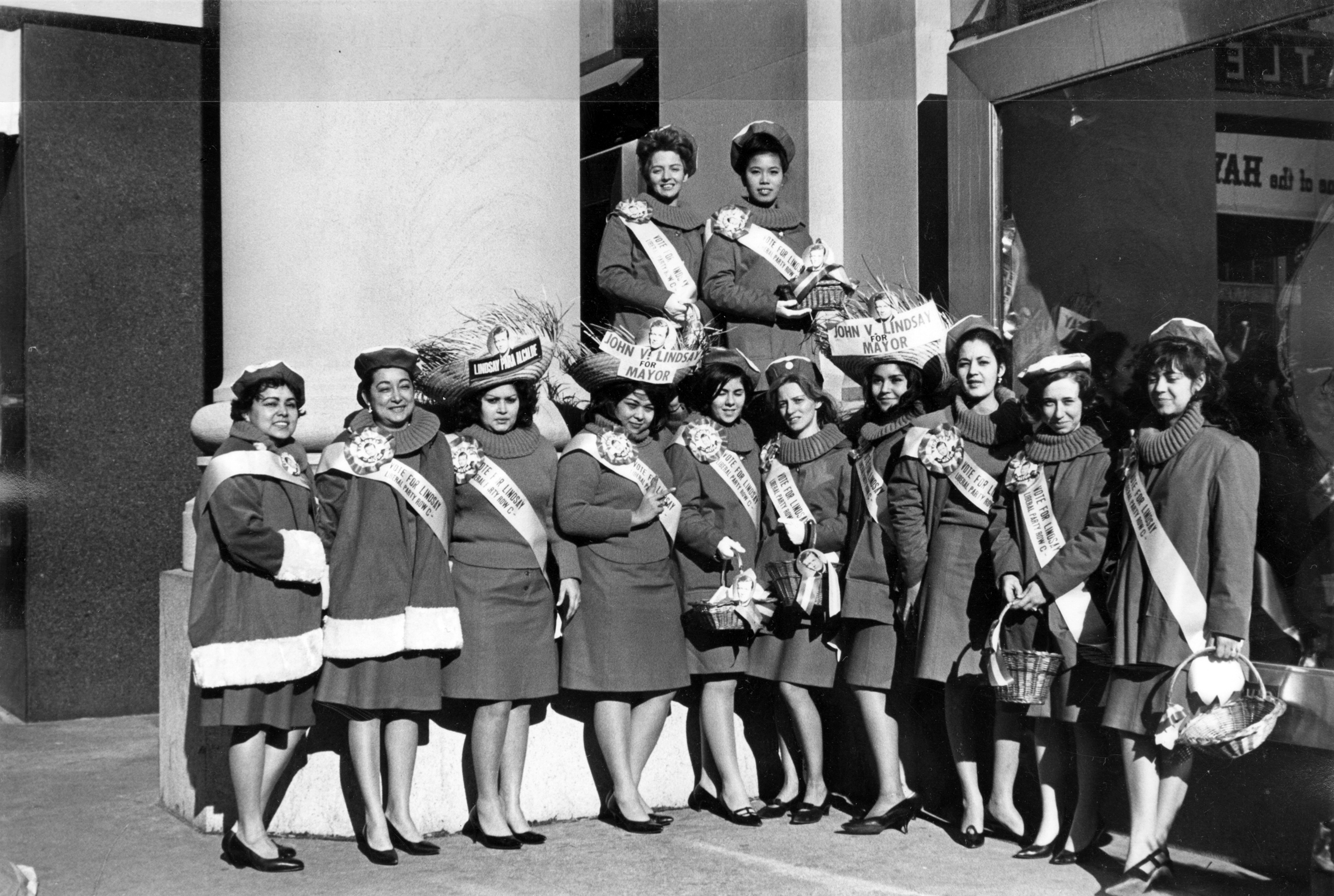 Kathy Andrade and a group of union women in matching coats and sashes holding campaign baskets and signs supporting John Lindsay for mayor, 1960s.