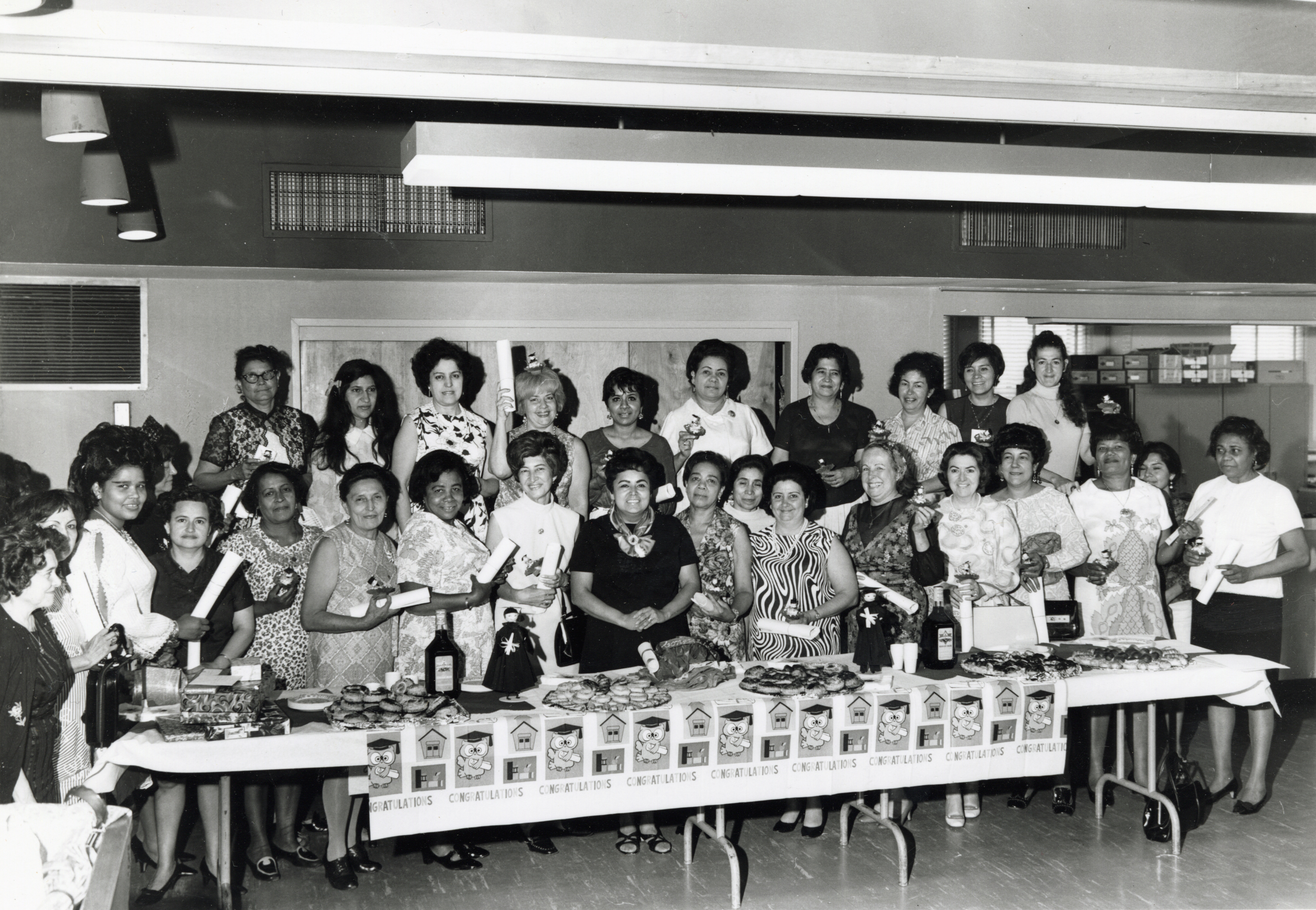 Kathy Andrade standing with a large group of women around a table decorated with food and graduation-themed décor, celebrating the completion of a citizenship class.