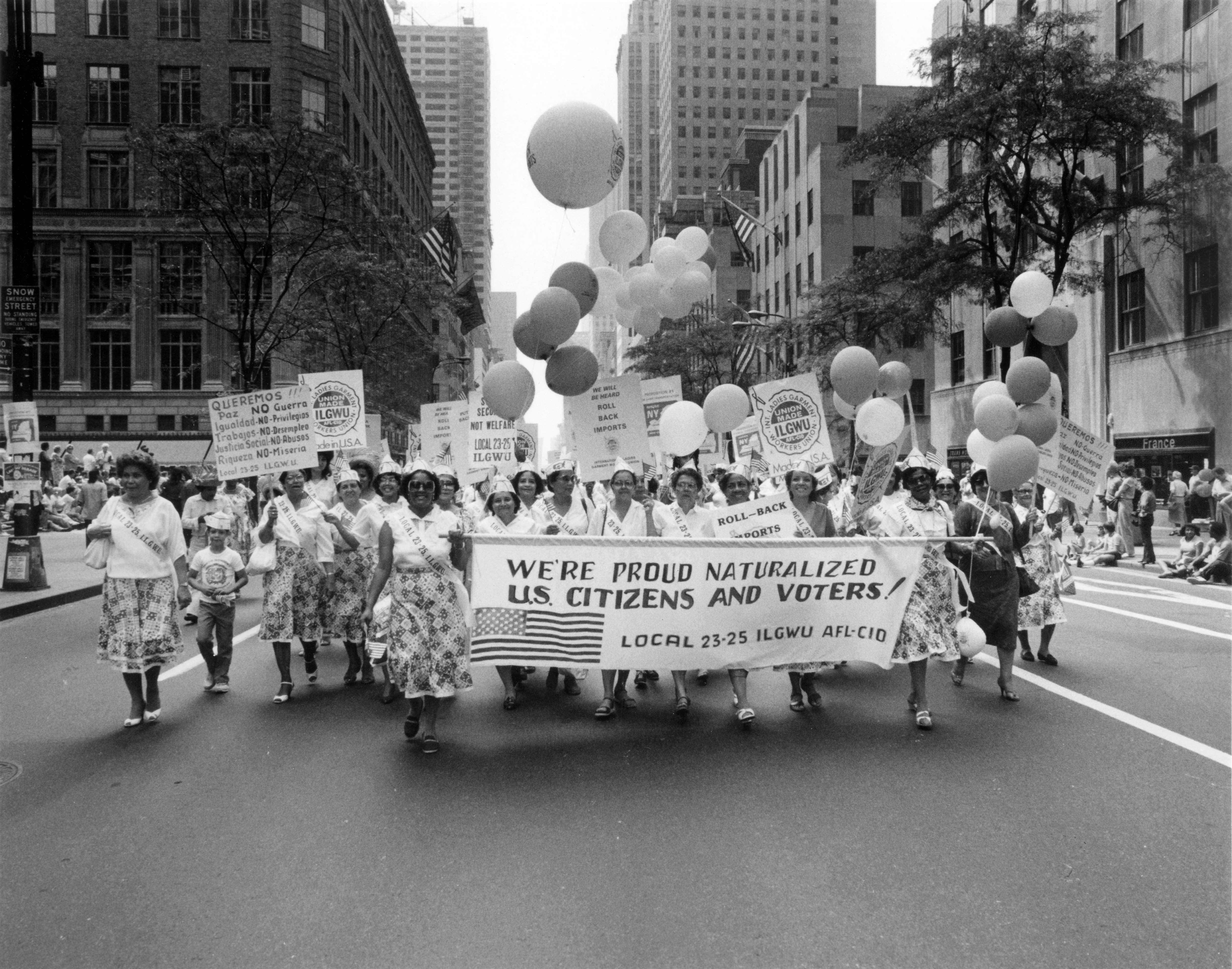 Union women in patterned skirts march in a Labor Day Parade, carrying balloons and a banner reading "We're proud naturalized U.S. citizens and voters! Local 23-25 ILGWU AFL-CIO," 1980s.